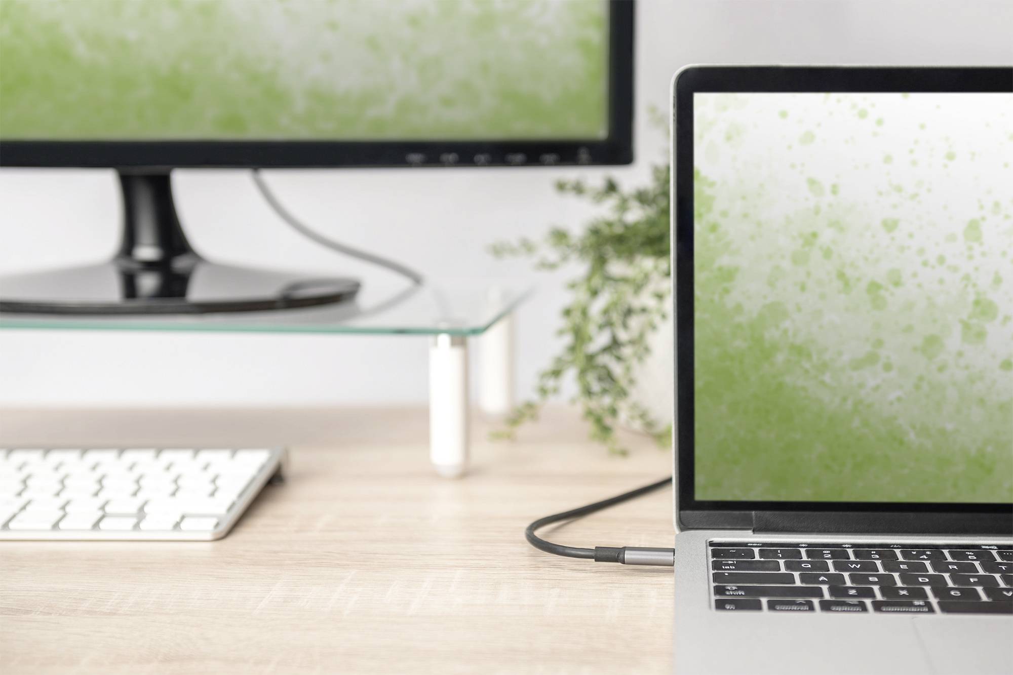 Ein Laptop und ein Monitor auf einem Schreibtisch, beide Bildschirme zeigen ein grünes Spritzmuster. Im Hintergrund eine Pflanze.