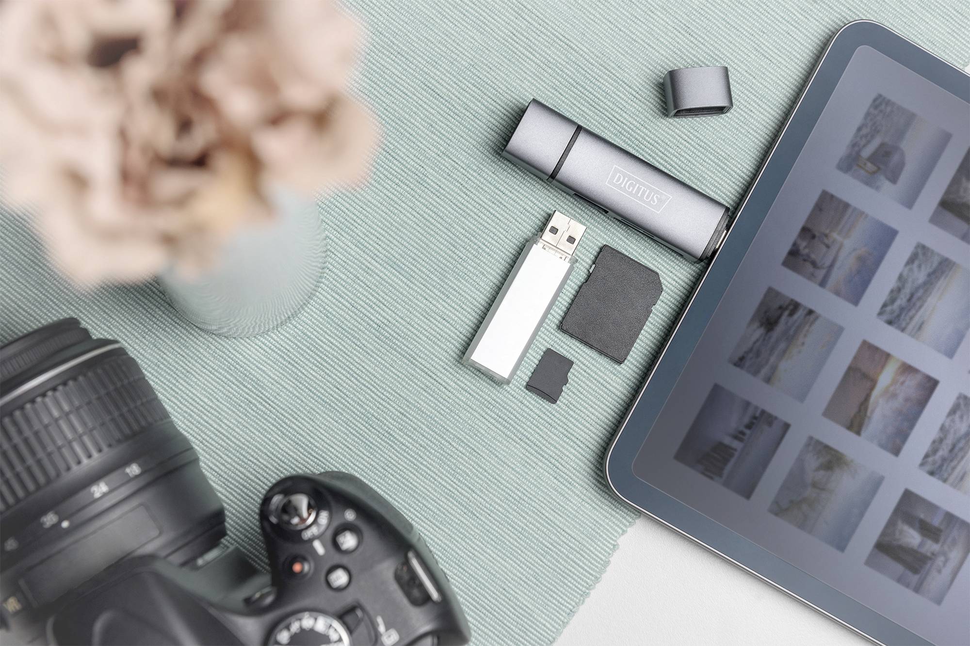 Eine Spiegelreflexkamera, ein Tablet mit Fotogalerie, ein USB-Laufwerk und SD-Karten auf einer türkisfarbenen Unterlage, daneben Blumen.