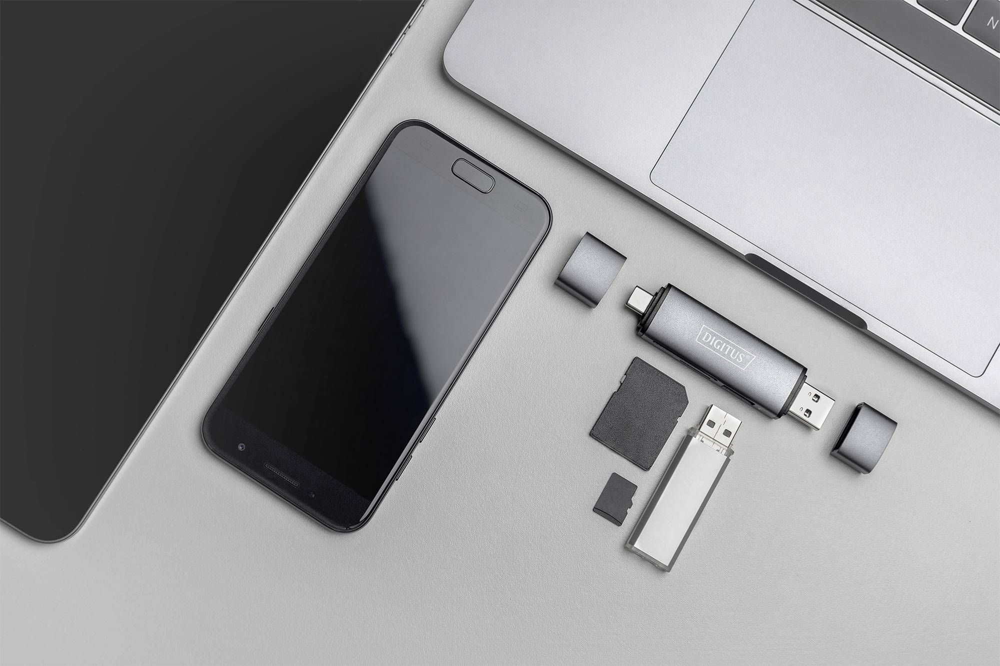 Smartphone, Laptop, USB-Stick, MicroSD und Adapter auf grauer Oberfläche. Technische Geräte für digitale Speicher- und Verbindungsaufgaben.