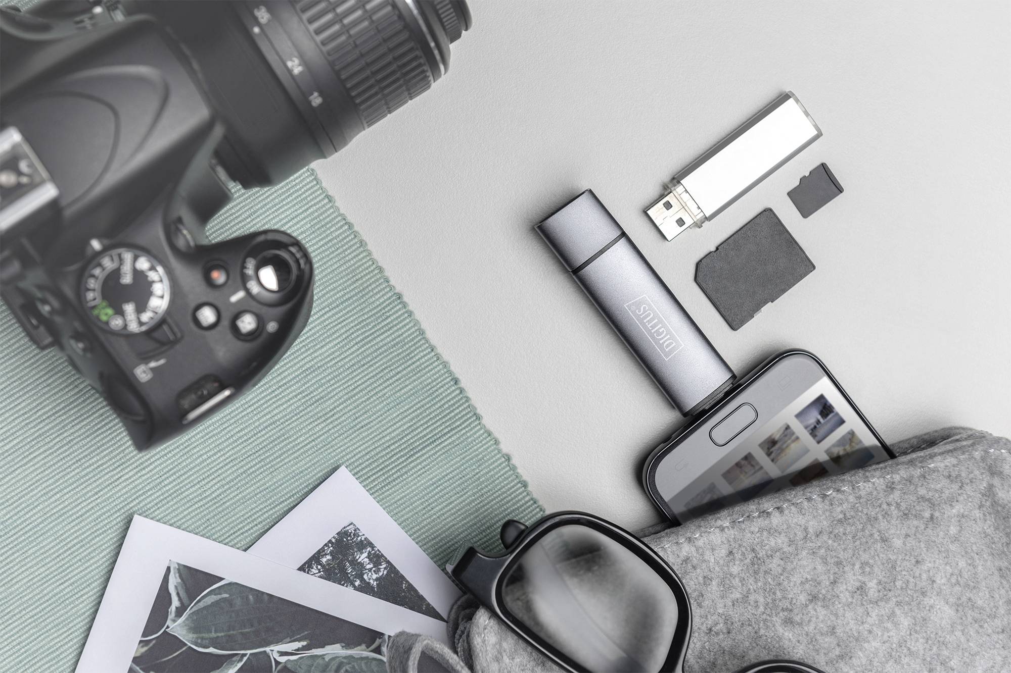 Kamera, USB-Stick, Smartphone, Brille und Fotos auf grauem Untergrund; repräsentiert digitale und analoge Fotografie.