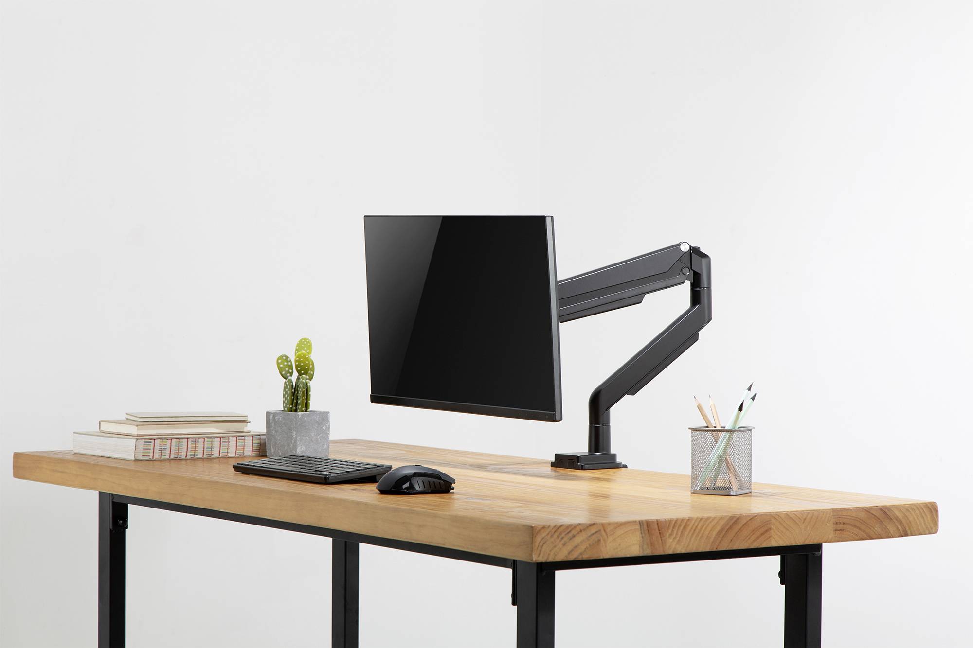 Ein moderner, aufgeräumter Schreibtisch mit Monitorarm, Monitor, Tastatur, Maus, Kaktuspflanze, Bücherstapel und Stiftehalter.