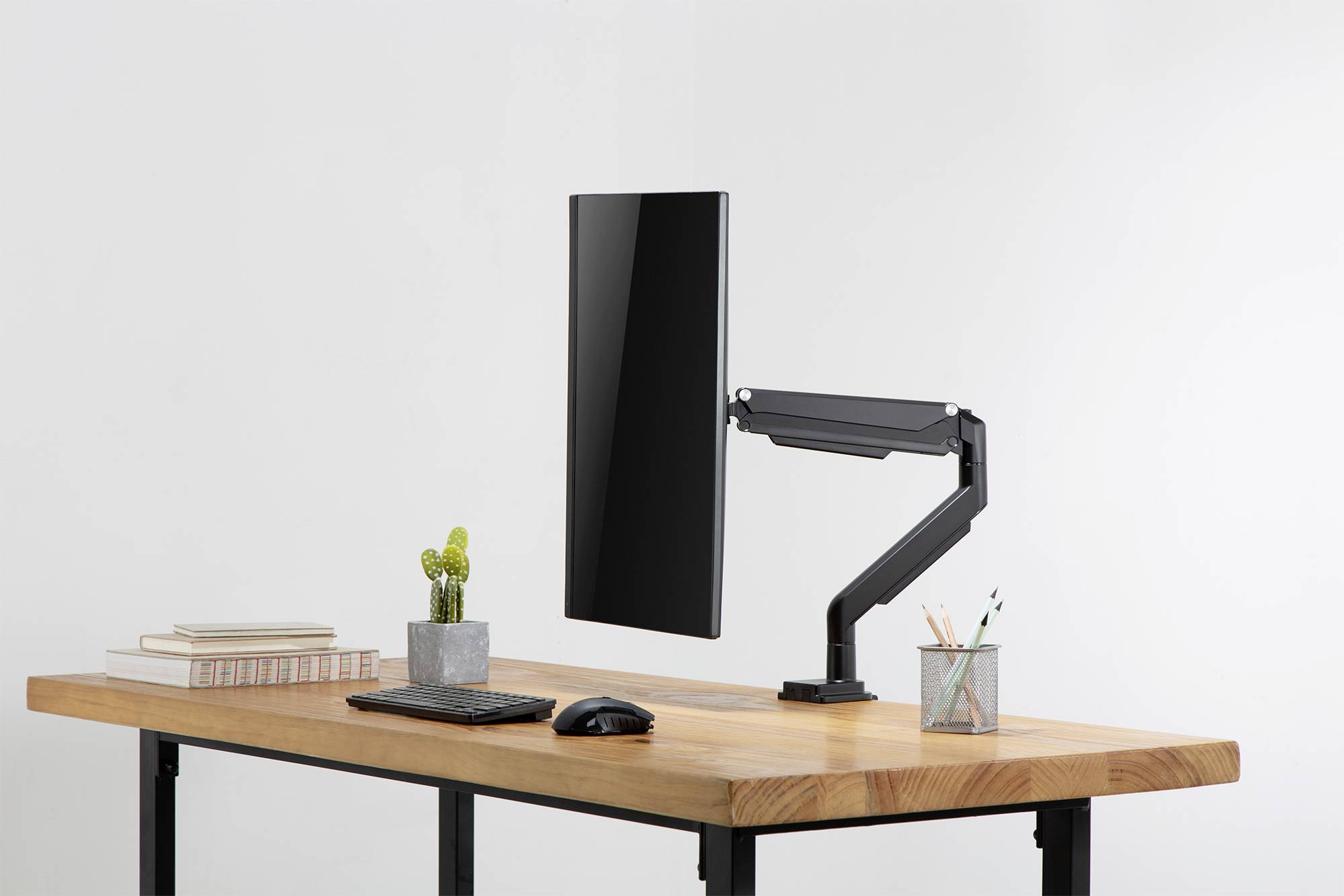 Ein moderner Schreibtisch mit einem Bildschirm an einem schwenkbaren Arm, einer Tastatur, einer Maus, Büchern, einem kleinen Kaktus und Schreibutensilien.