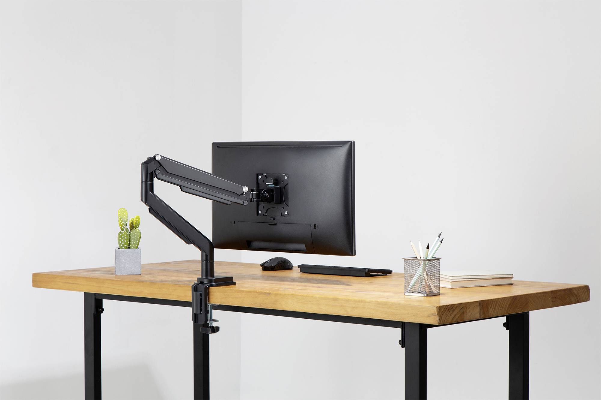 Ein moderner Schreibtisch mit Holzplatte und schwarzem Metallgestell. Darauf stehen ein Monitor mit Gelenkarm, ein kleiner Kaktus und Stifte.