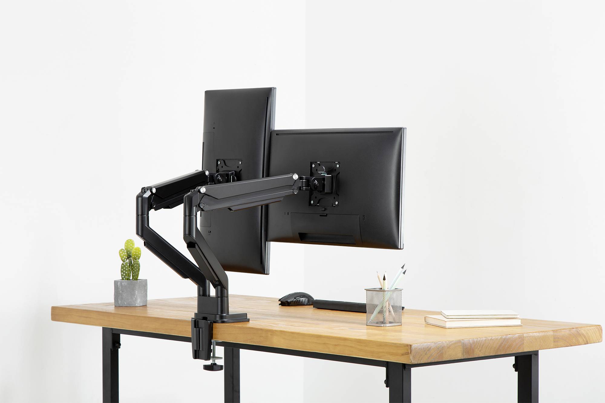 Zwei schwarze Monitore auf einem höhenverstellbaren, schwenkbaren Monitorarm, montiert auf einem Holztisch. Die Schreibtischecke hat Pflanzen und Schreibutensilien.