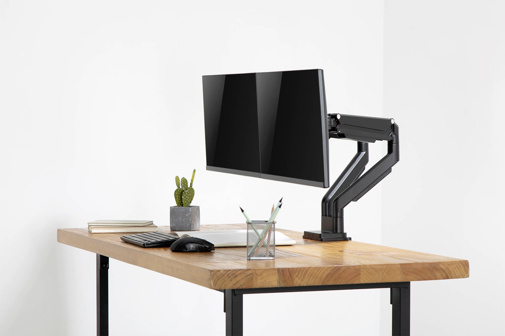 Ein moderner Schreibtisch mit Monitor an einem verstellbaren Arm, daneben befinden sich Tastatur, Maus, Notizbuch, Stiftehalter und ein Kaktus.