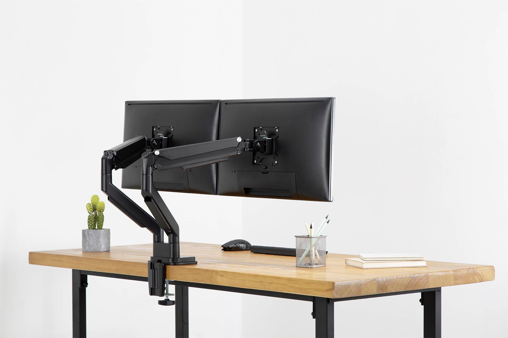 Zwei nebeneinander montierte Computermonitore auf einem verstellbaren Arm stehen auf einem Holzschreibtisch, daneben Pflanzen und Bürobedarf.