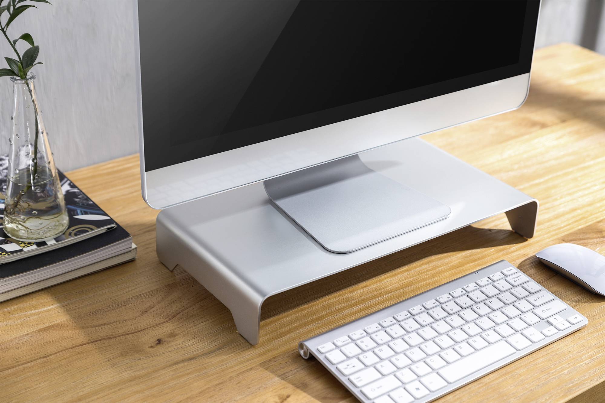 Ein Computerbildschirm auf einem weißen Standfuß, mit Tastatur und Maus auf einem Holztisch. Im Hintergrund sind Bücher und eine Vase.