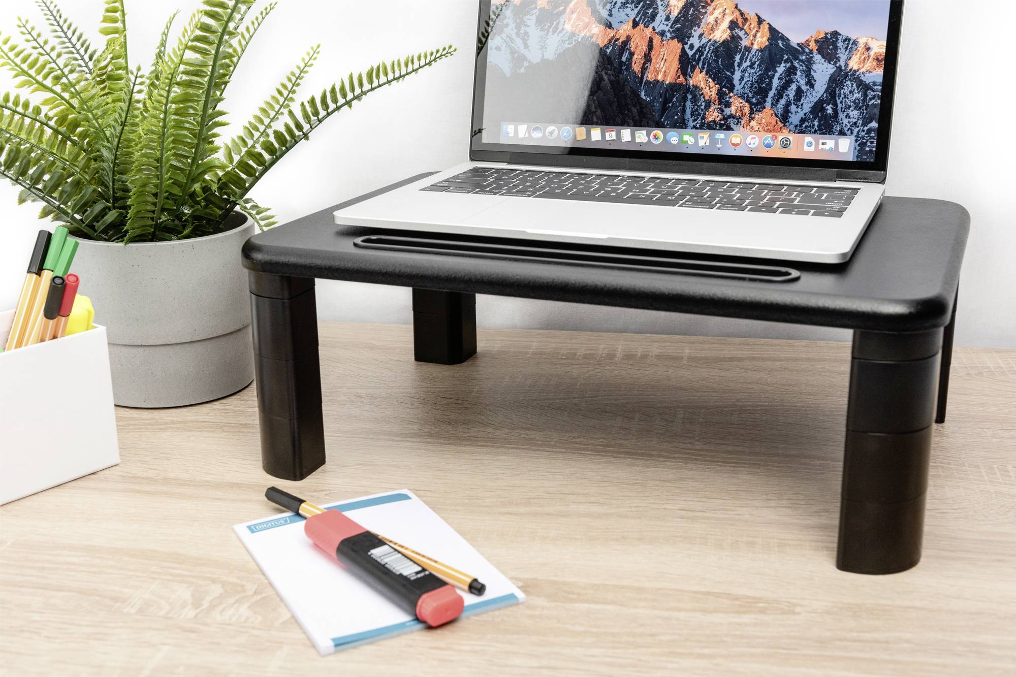 Ein Laptop steht auf einem schwarzen Tischständer. Neben dem Tisch befinden sich ein Topf mit einer Pflanze und Büroartikel.