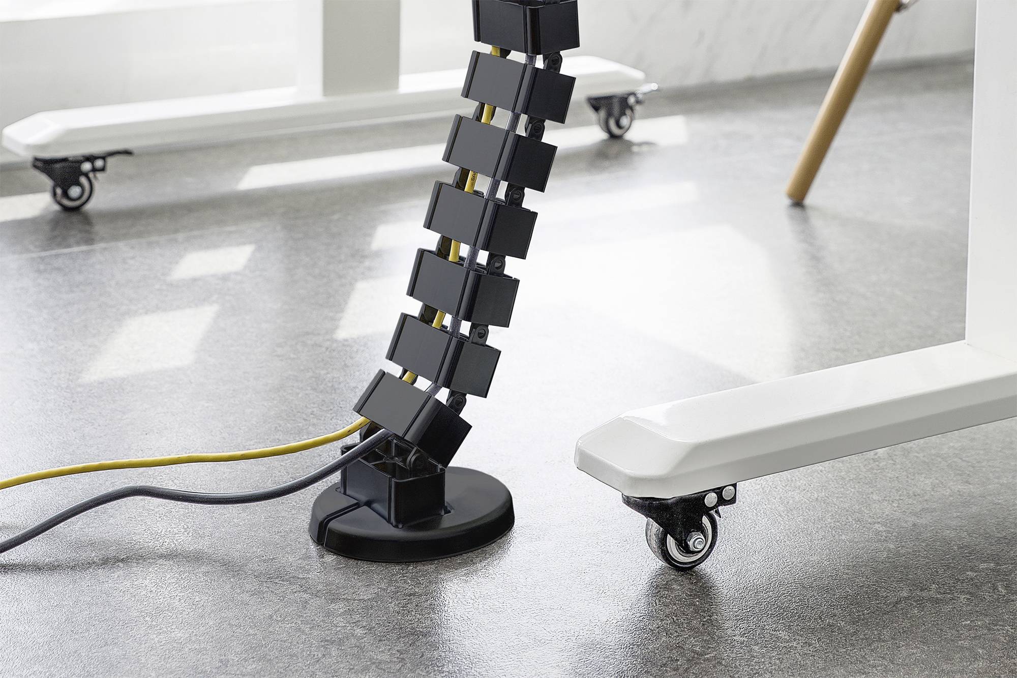 Eine flexible Kabelkette in Schwarz führt von einem Tisch auf den Boden. Kabel sind darin ordentlich verstaut. Tischbeine auf Rollen sichtbar.