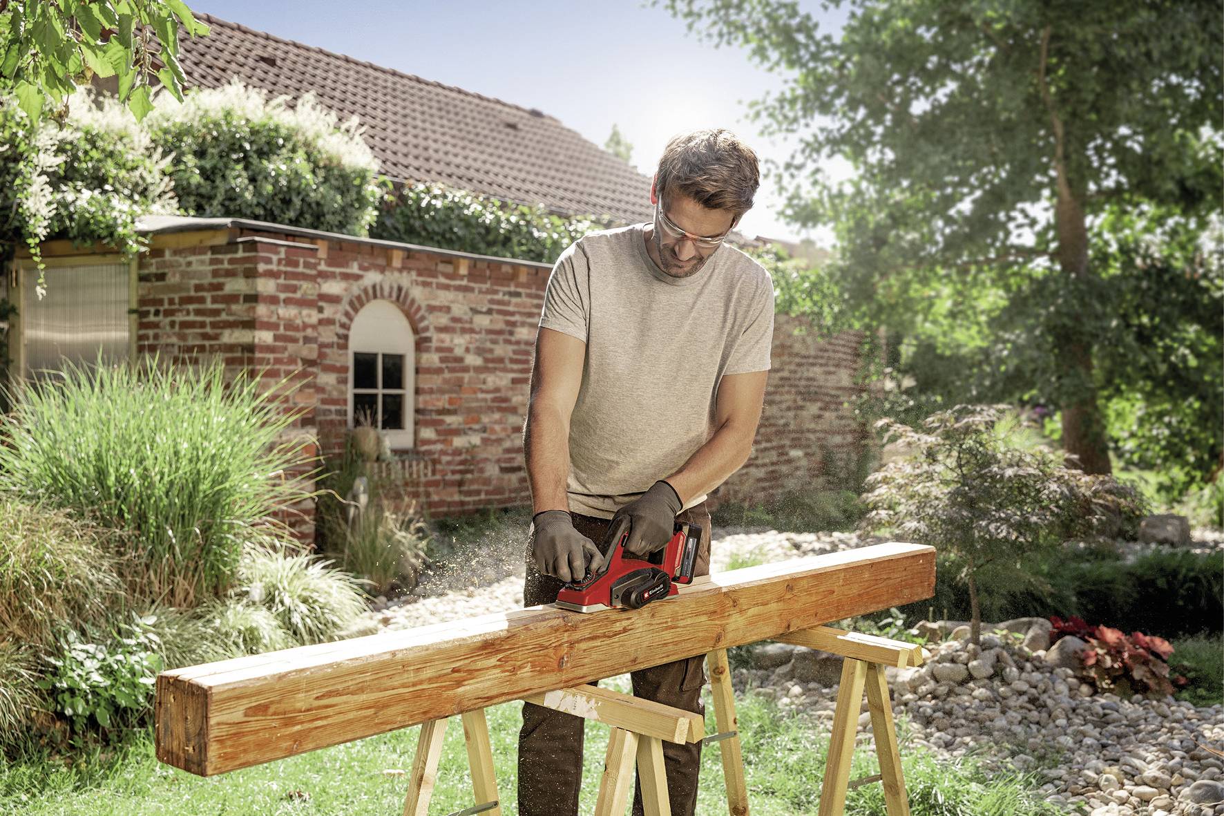 Ein Mann schleift draußen einen Holzbalken auf zwei Böcken in einem sonnigen Garten. Im Hintergrund ist ein Backsteinhaus zu sehen.
