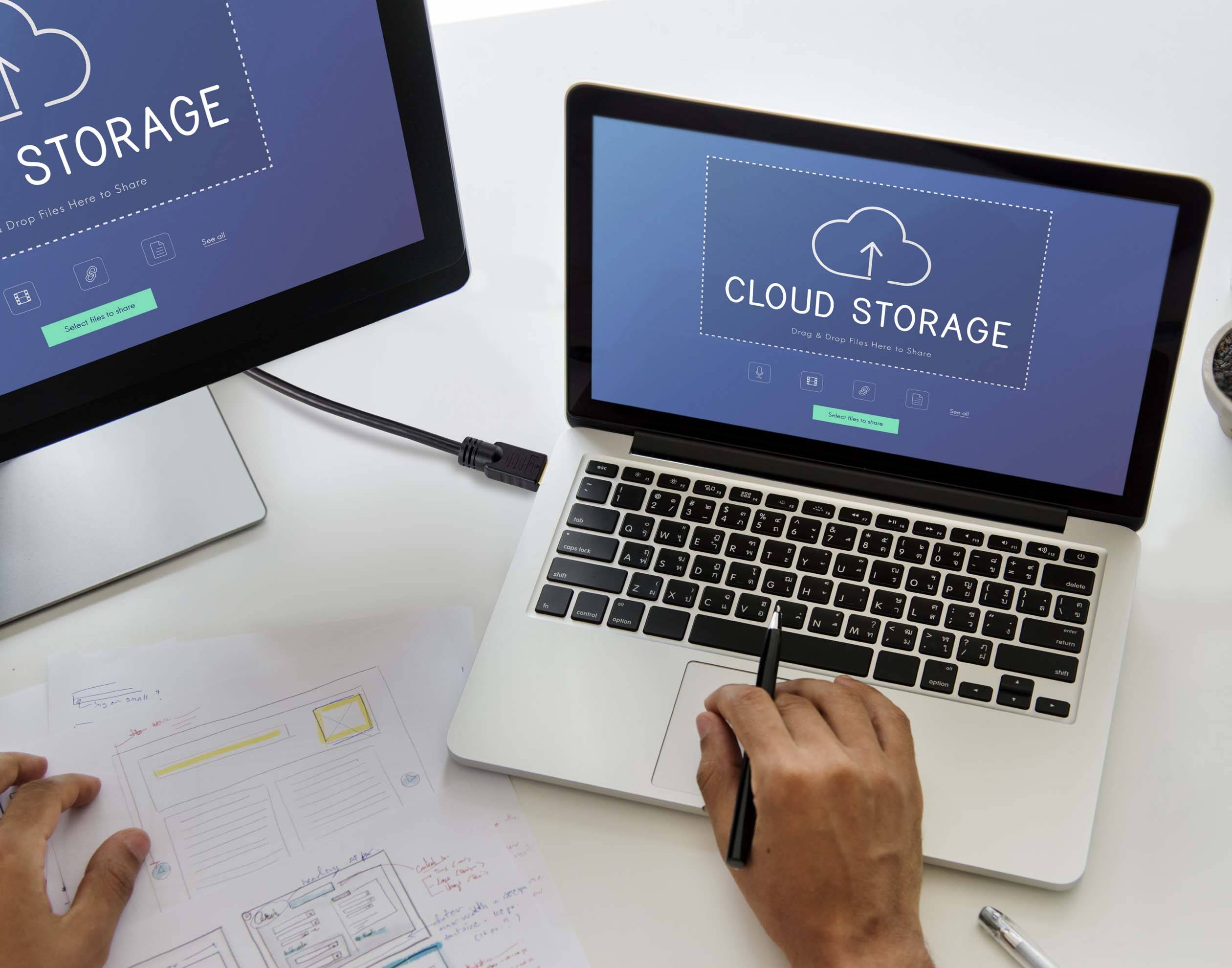 Zwei Bildschirme zeigen 'Cloud Storage'-Seiten. Eine Person arbeitet an einem Laptop und schreibt auf Papier. Es geht um Online-Datenspeicherung.