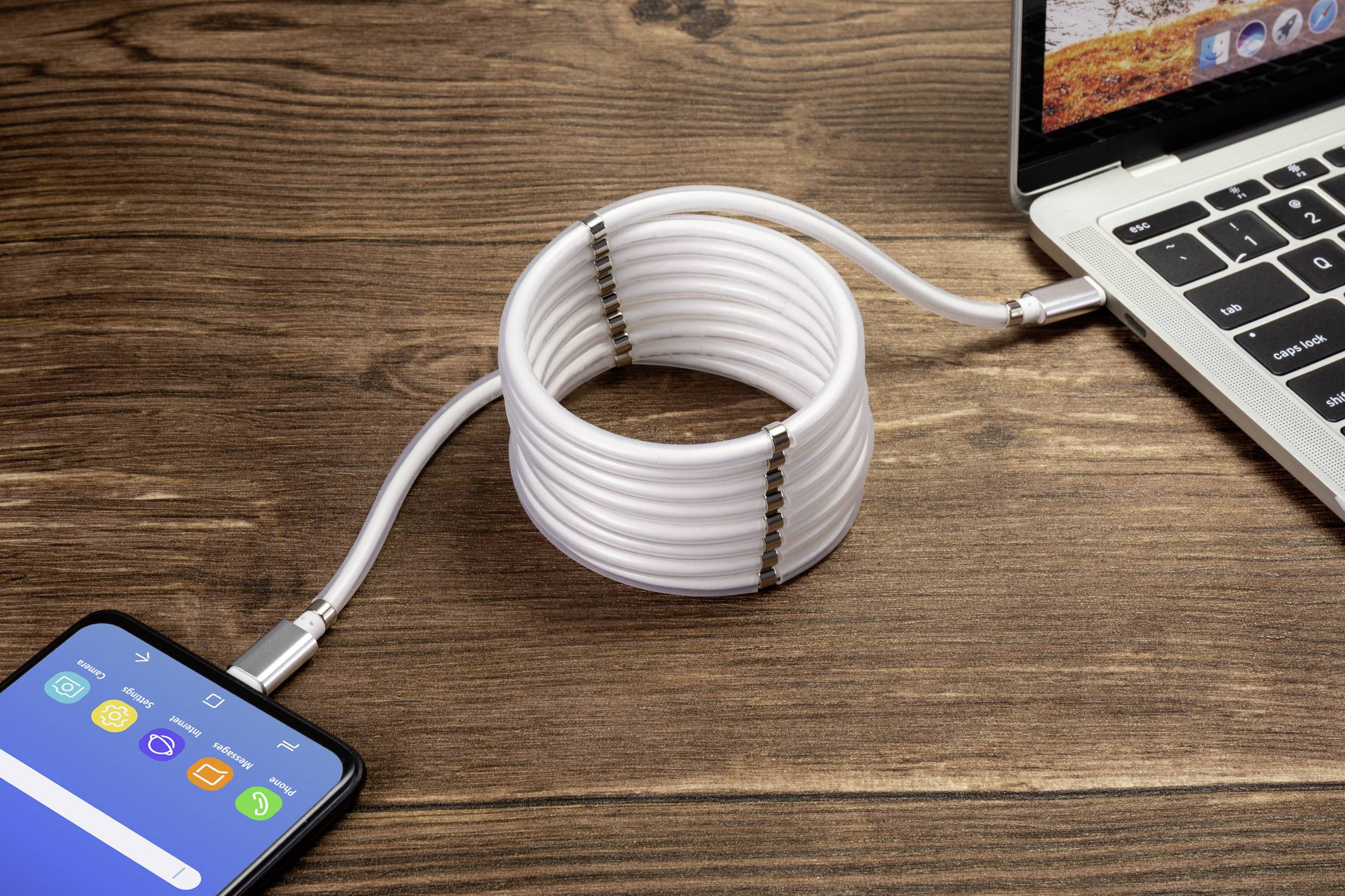 Ein Smartphone und ein Laptop sind durch ein weißes, spiralförmig gewickeltes Kabel auf einem hölzernen Tisch verbunden.