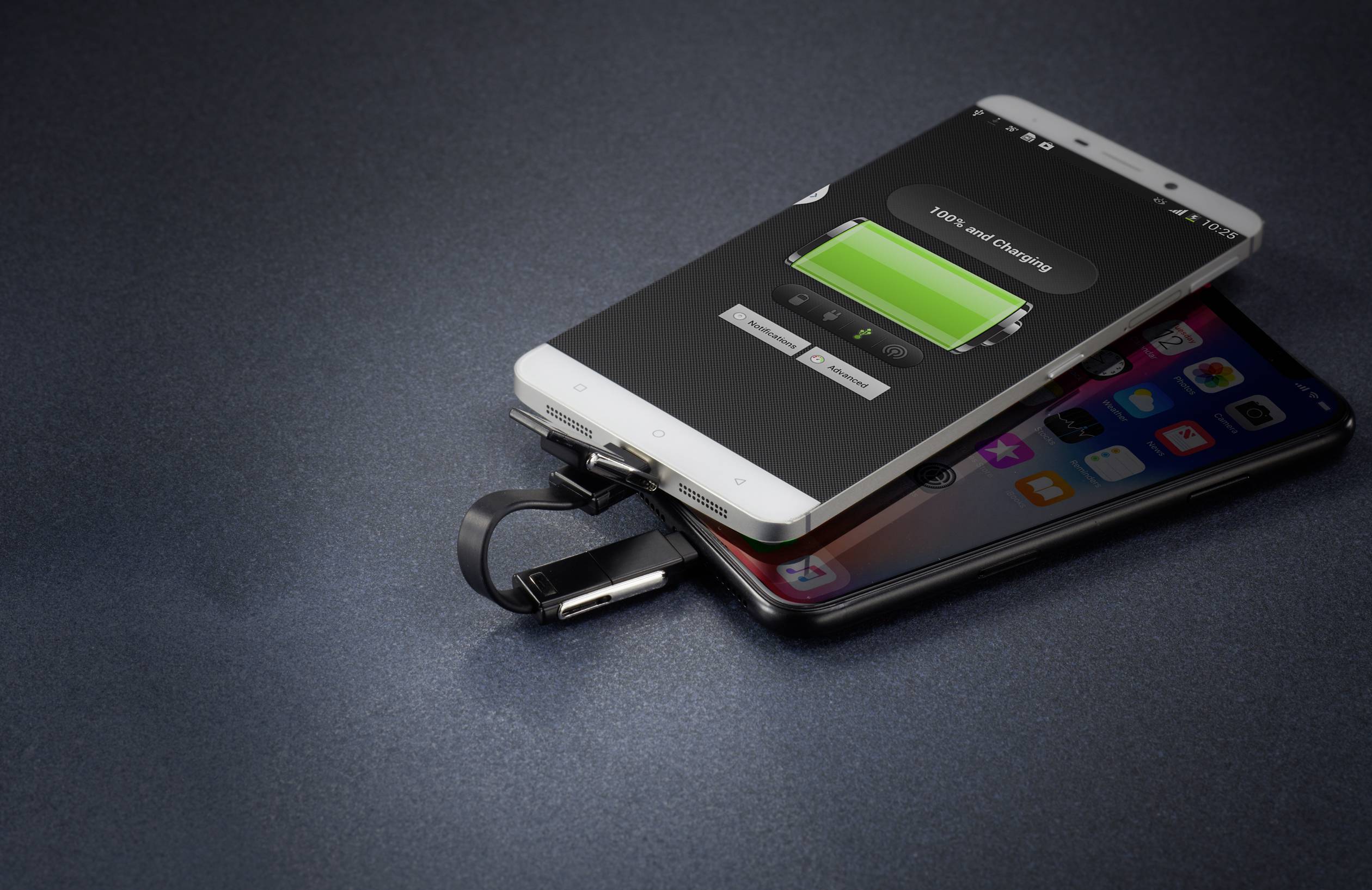 Zwei Smartphones übereinander, eines mit Ladeanzeige und angeschlossenem USB-Kabel, auf einer dunklen Oberfläche liegend.