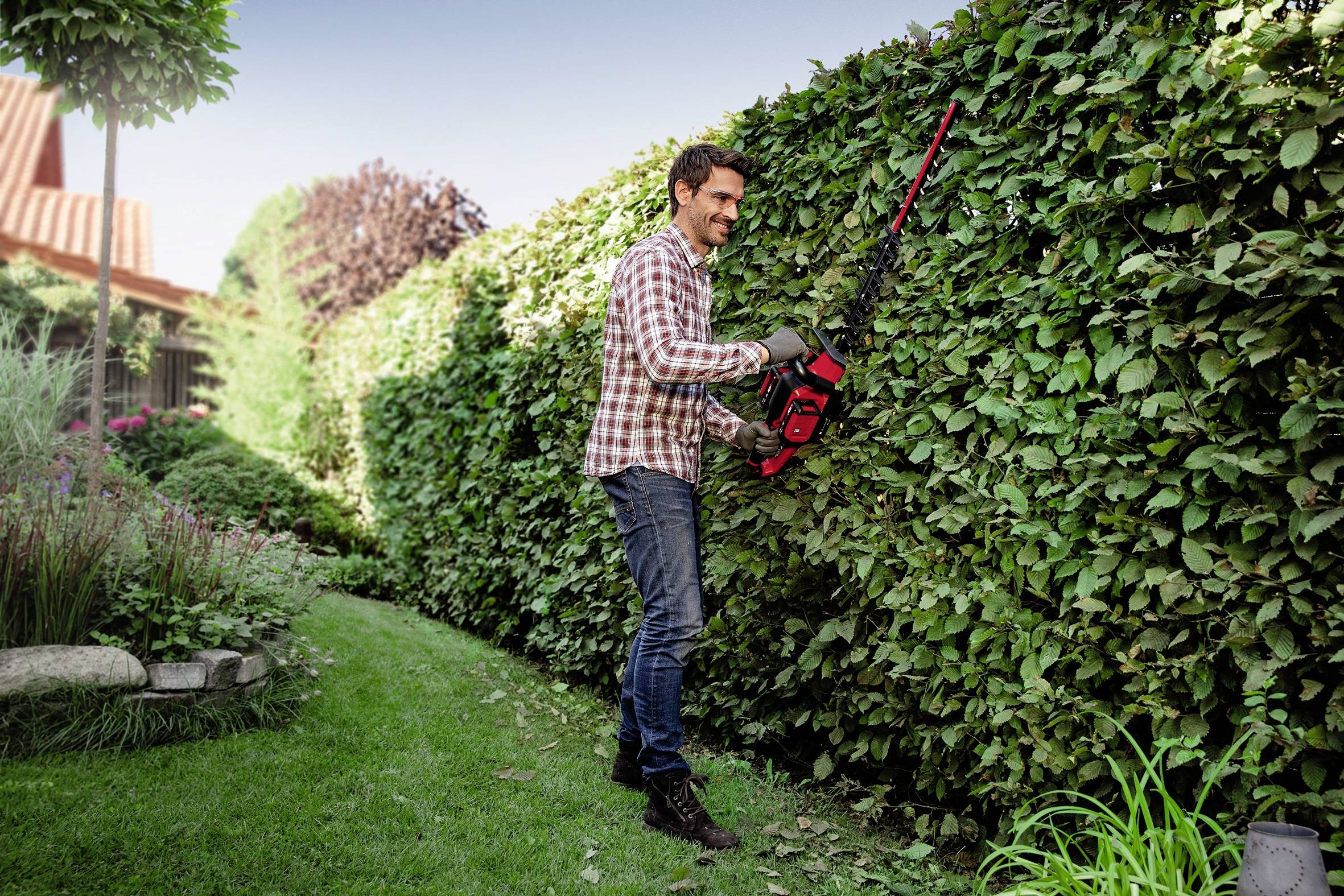 Mann schneidet Hecke mit elektrischer Heckenschere im Garten. Er trägt ein kariertes Hemd und Jeans, umgeben von Pflanzen und einem Haus.