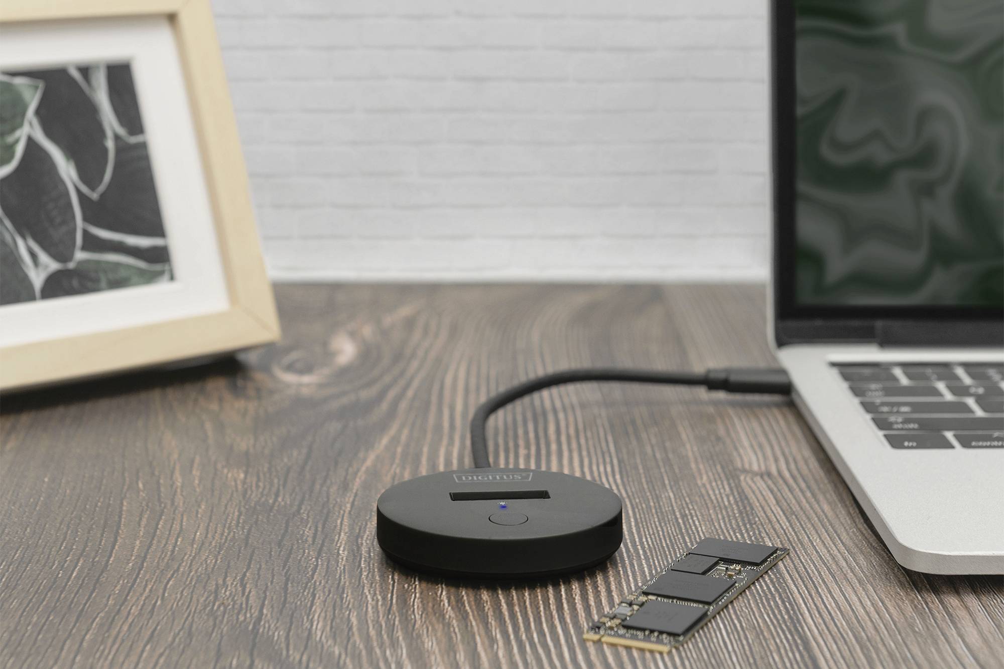 Ein runder Adapter mit Kabel verbindet einen Laptop auf einem Holztisch. Ein Bilderrahmen steht im Hintergrund, daneben liegt ein Speichermodul.