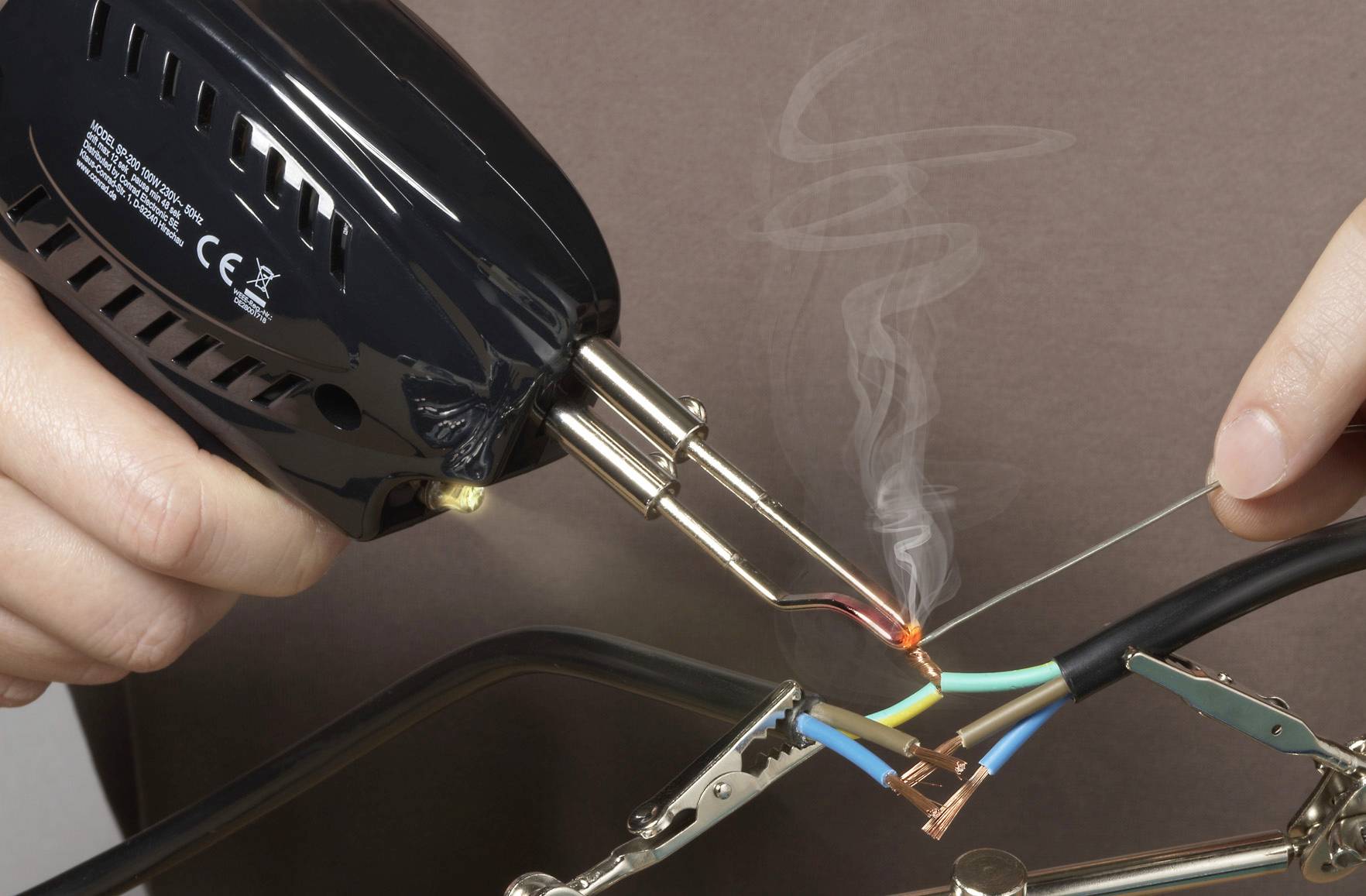 Eine Person lötet zwei Drähte. Rauch steigt von der Lötstelle auf. Die Drähte sind durch farbige Isolierungen unterscheidbar.