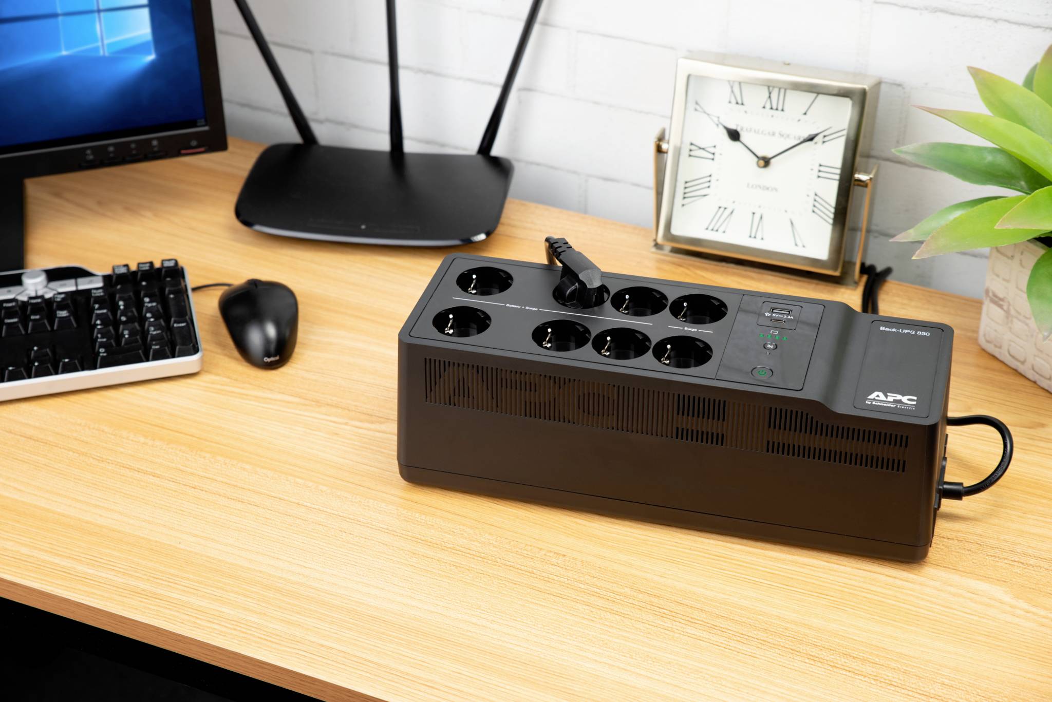 Auf einem Schreibtisch steht ein APC-Überspannungsschutz mit mehreren Steckdosen. Daneben befinden sich ein Monitor, eine Tastatur, eine Maus und eine Uhr.