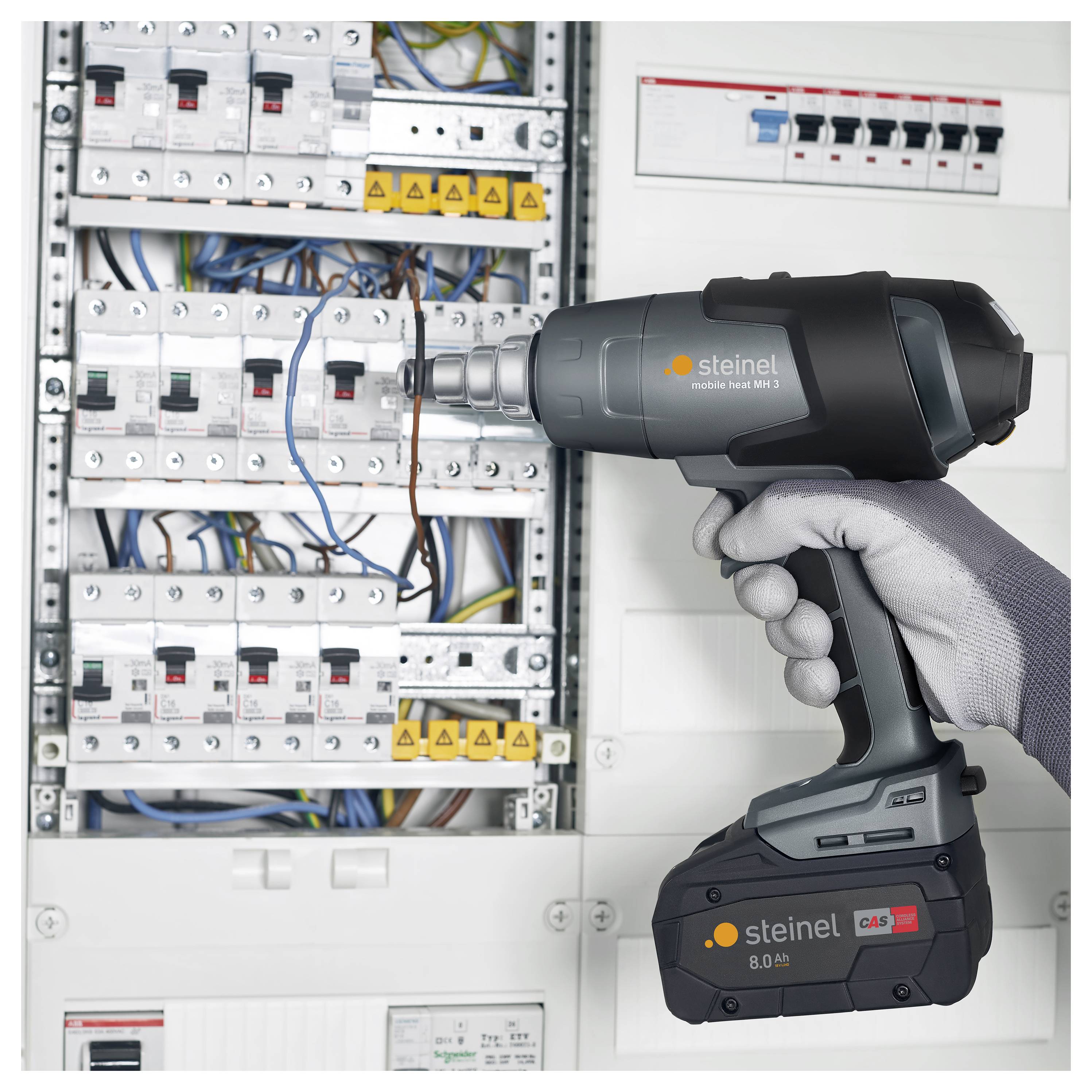 Eine behandschuhte Hand hält eine kabellose Heißluftpistole, die auf ein offenes Elektroschaltfeld mit Leistungsschaltern und freiliegenden Leitungen gerichtet ist.