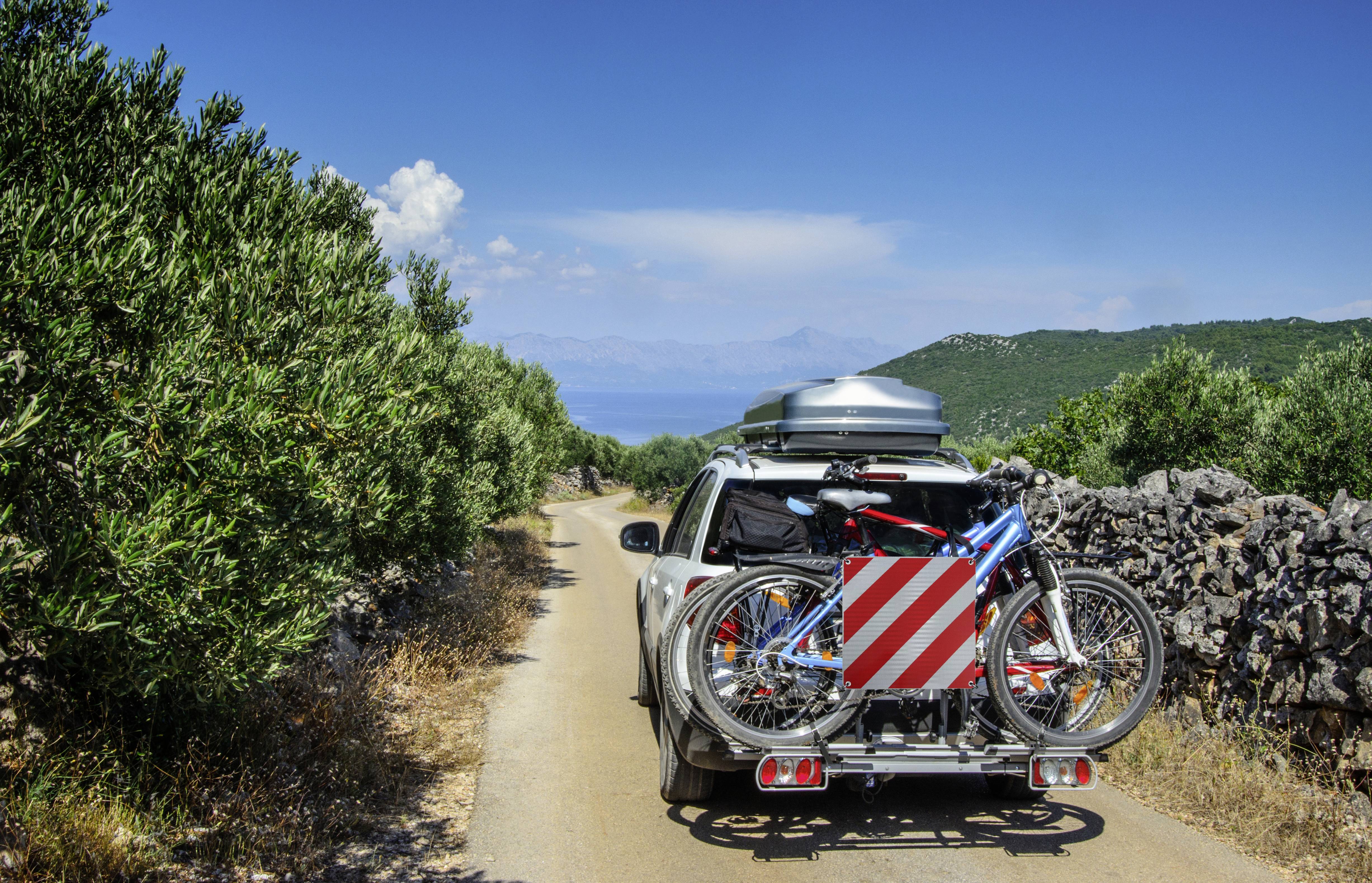 Ein Auto fährt auf einer schmalen Straße durch eine grüne Landschaft, mit zwei Fahrrädern am Heckträger und einer Gepäckbox auf dem Dach.