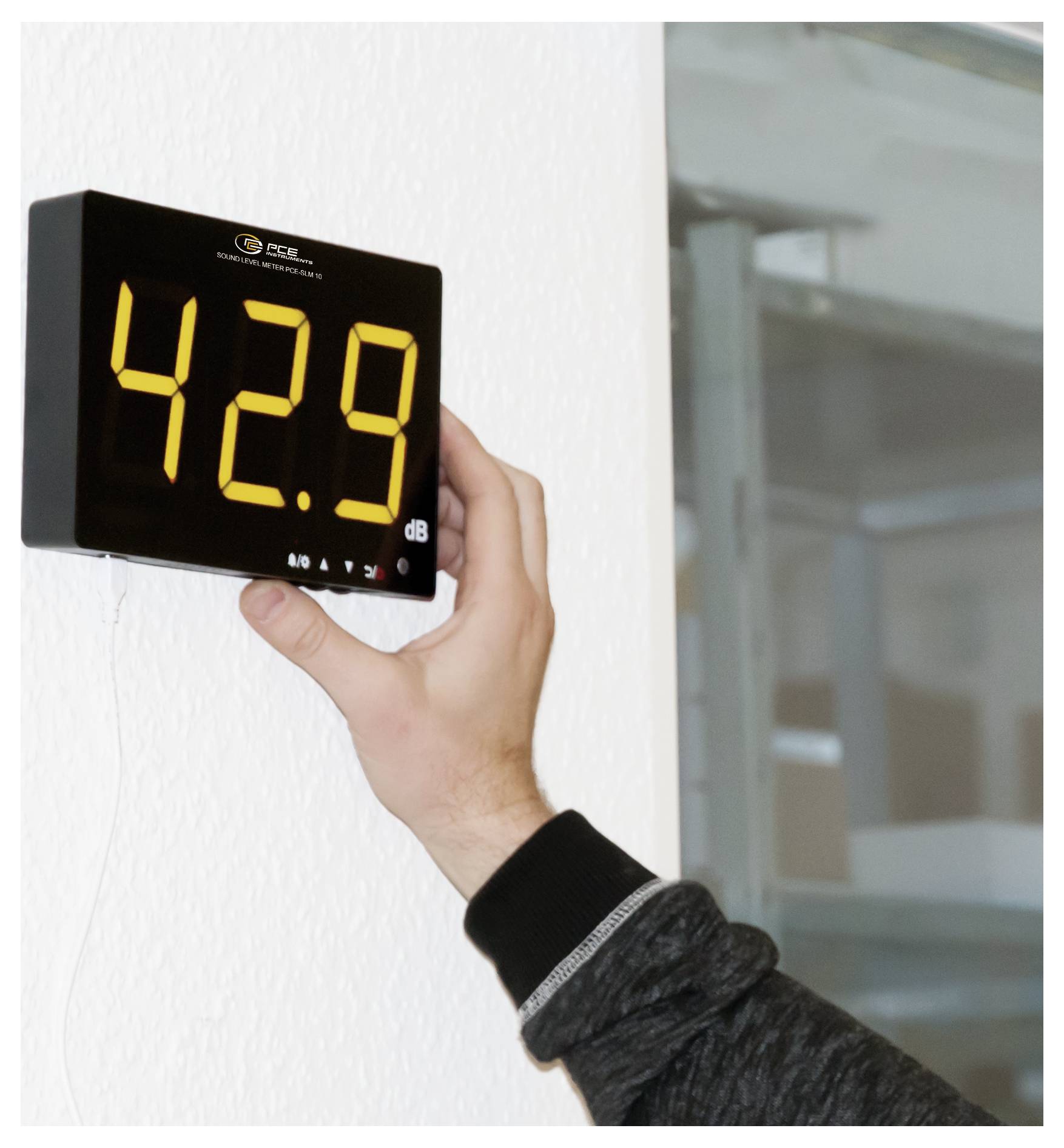 Hand hält ein digitales Schallpegelmessgerät an einer Wand an, das 42,9 dB anzeigt.