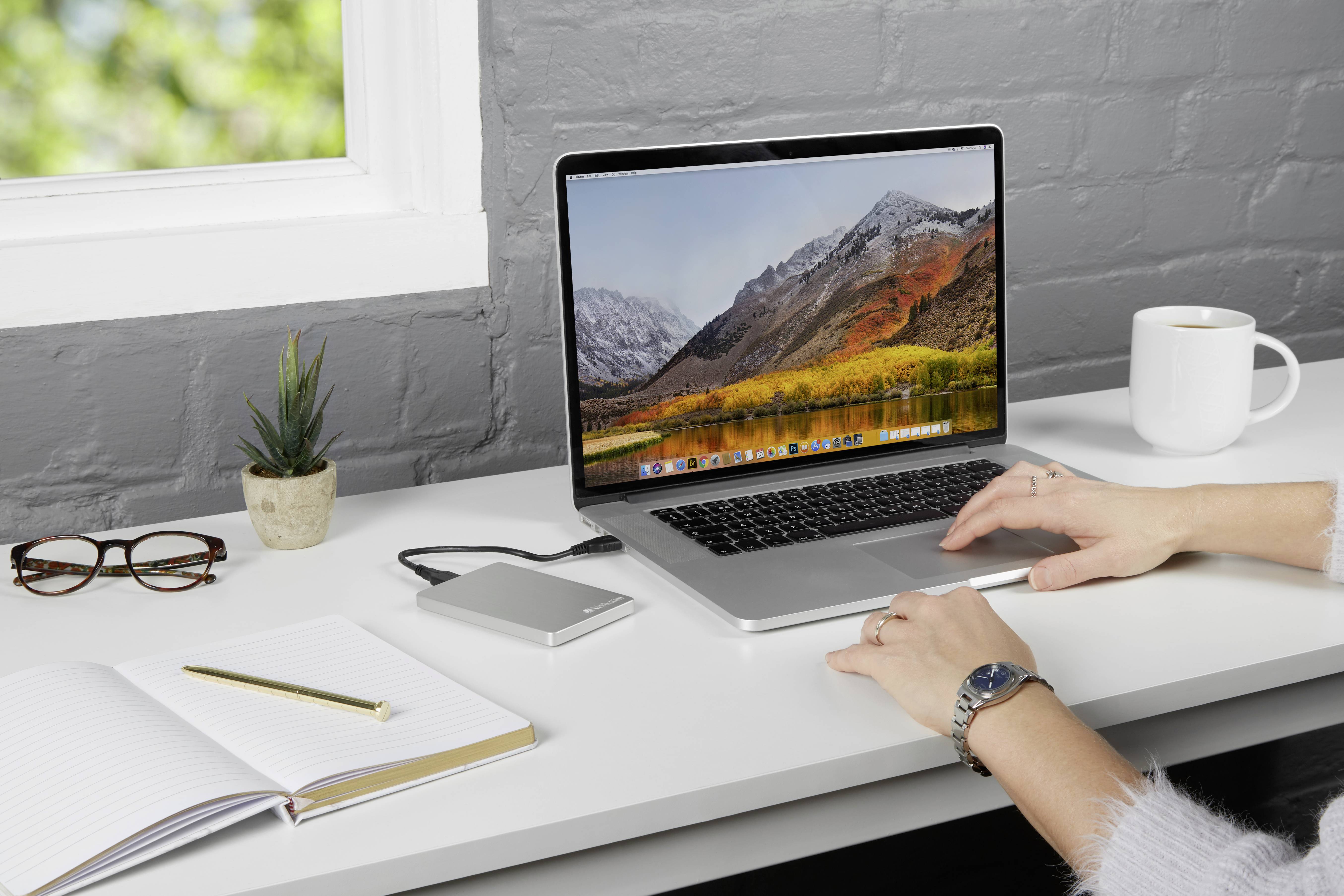 Eine Person arbeitet an einem Laptop auf einem Schreibtisch mit Notizbuch, Brille, externer Festplatte, Pflanze und Kaffeetasse.