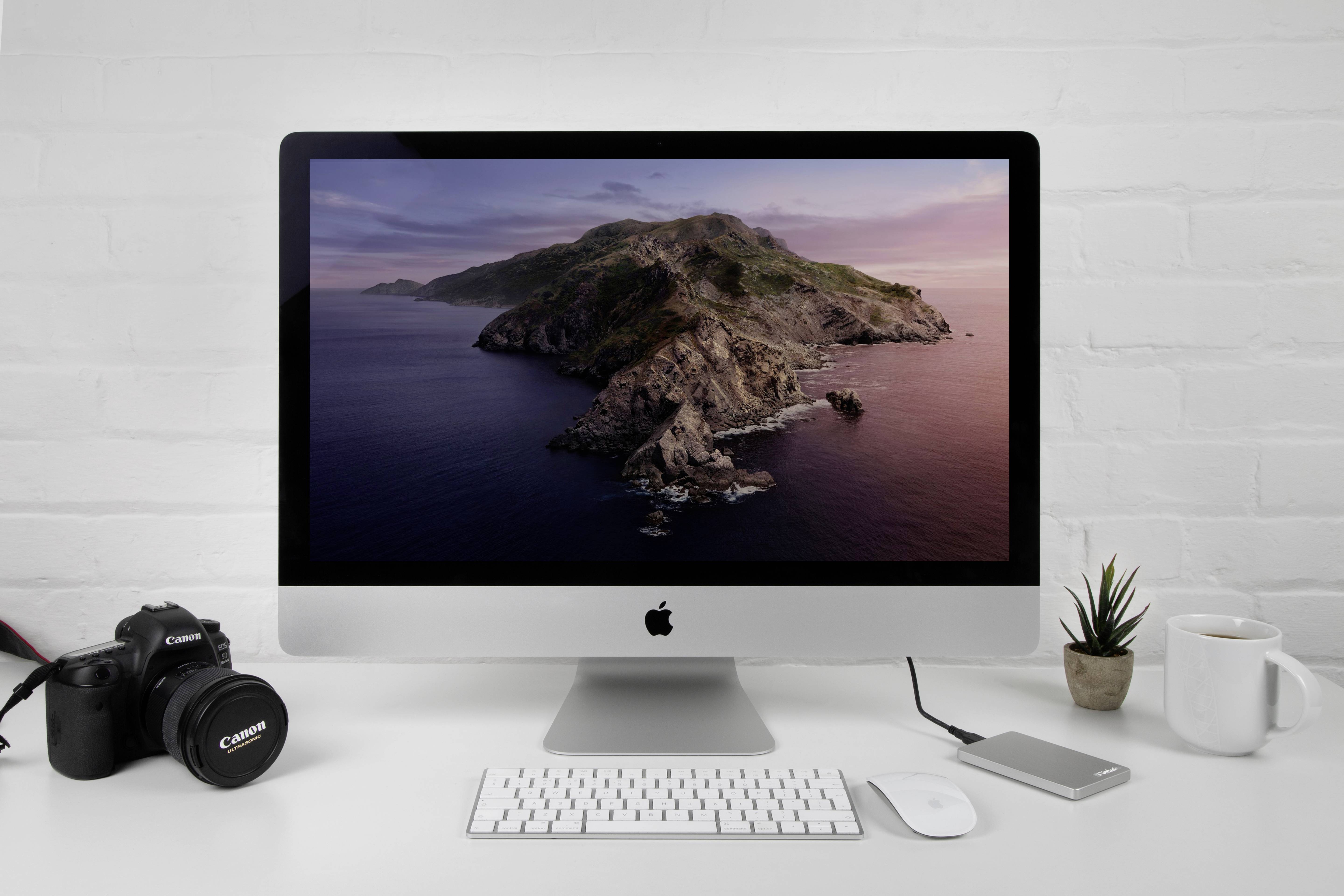 Ein Schreibtisch mit einem Apple-Computer, der ein Küstenbild anzeigt, daneben eine Kamera, eine externe Festplatte und eine Tasse.