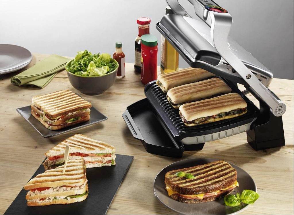 Ein Tisch mit einem Grillgerät, das mehrere Sandwiches toastet. Daneben stehen Teller mit fertigen Sandwiches und Schüsseln mit Salat.