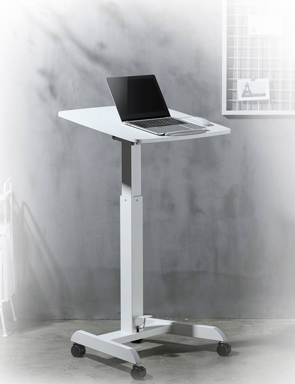 Ein höhenverstellbarer weißer Tisch mit Rollen, auf dem ein geöffneter Laptop steht, vor einer grauen Wand und einem weißen Gitterbrett.