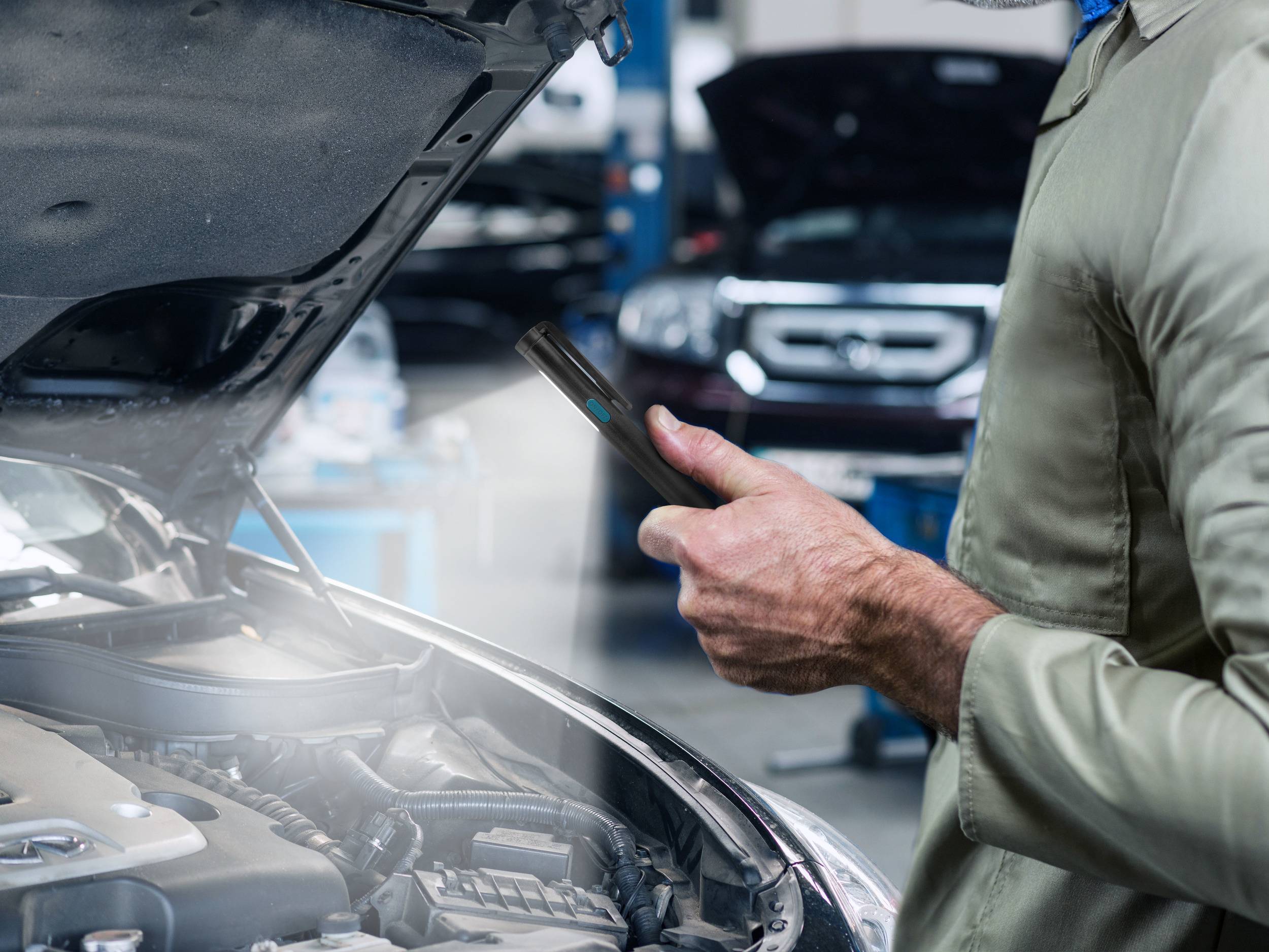 Ein Mechaniker steht neben einem offenen Auto und überprüft den Motor mit einem Smartphone. Im Hintergrund sind weitere Fahrzeuge.