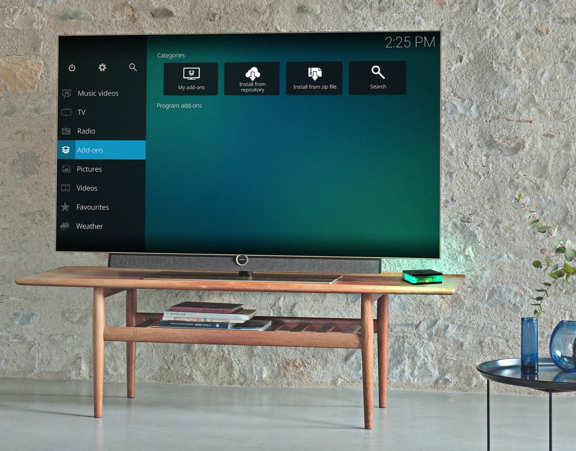 Smart-TV auf Holztisch, zeigt Menübildschirm mit Kategorien wie Musikvideos, Bilder und Add-ons. Uhrzeit: 14:25.