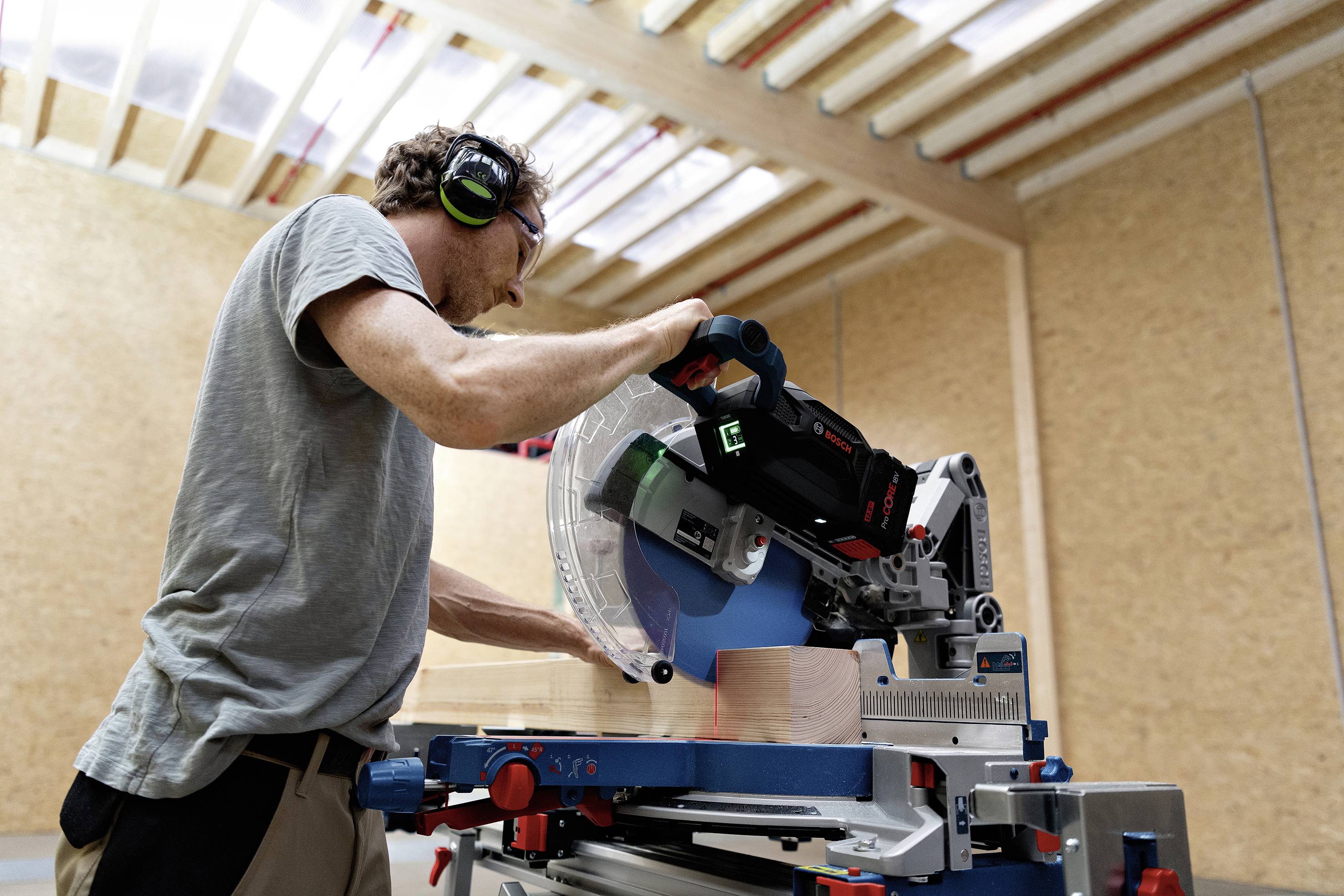 Ein Mann arbeitet in einer Werkstatt mit Kappsäge, schneidet Holz entlang einer markierten Linie. Er trägt Gehörschutz.
