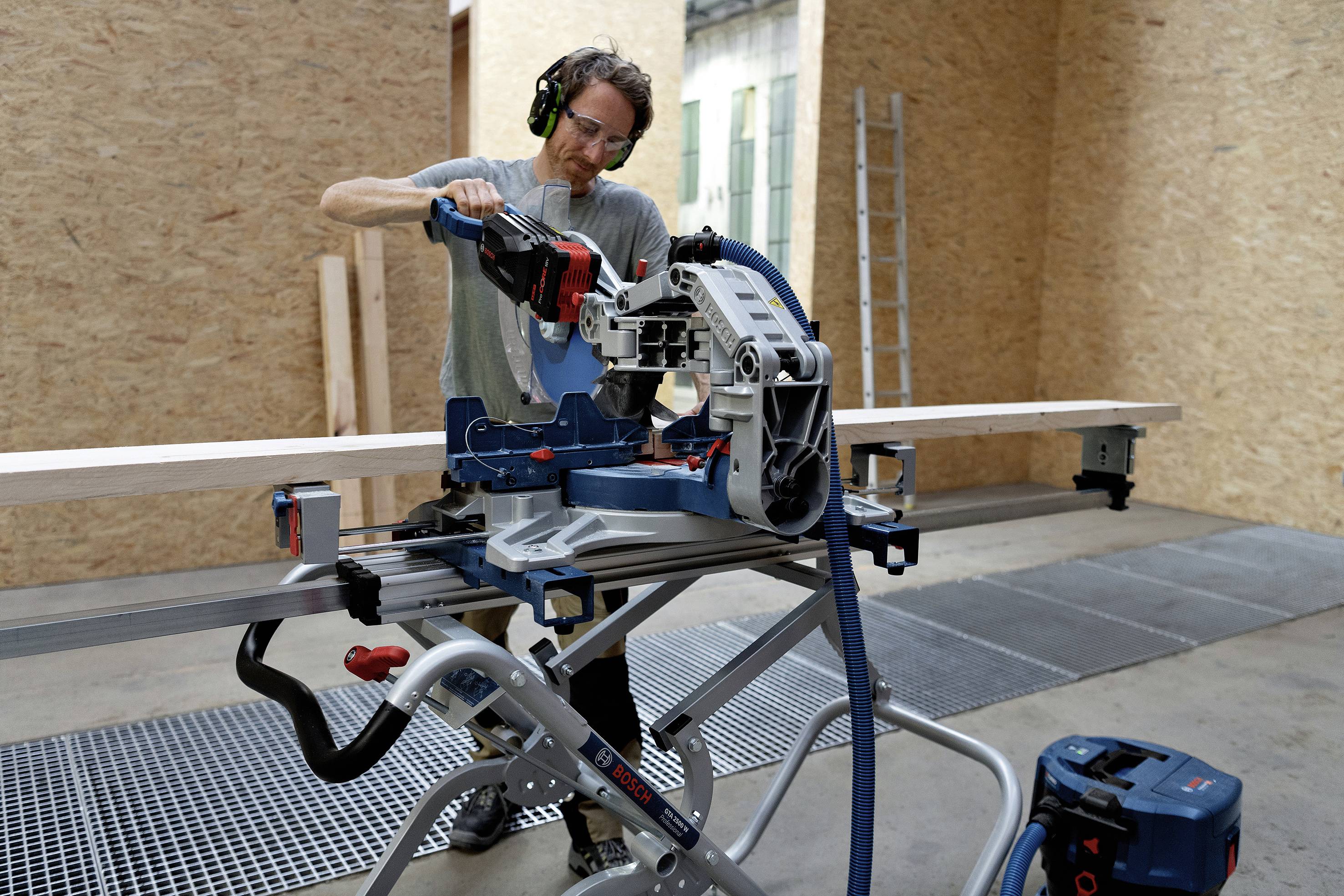 Ein Mann mit Gehörschutz schneidet Holz mit einer elektrischen Gehrungssäge in einer Werkstatt. Im Hintergrund eine Leiter.