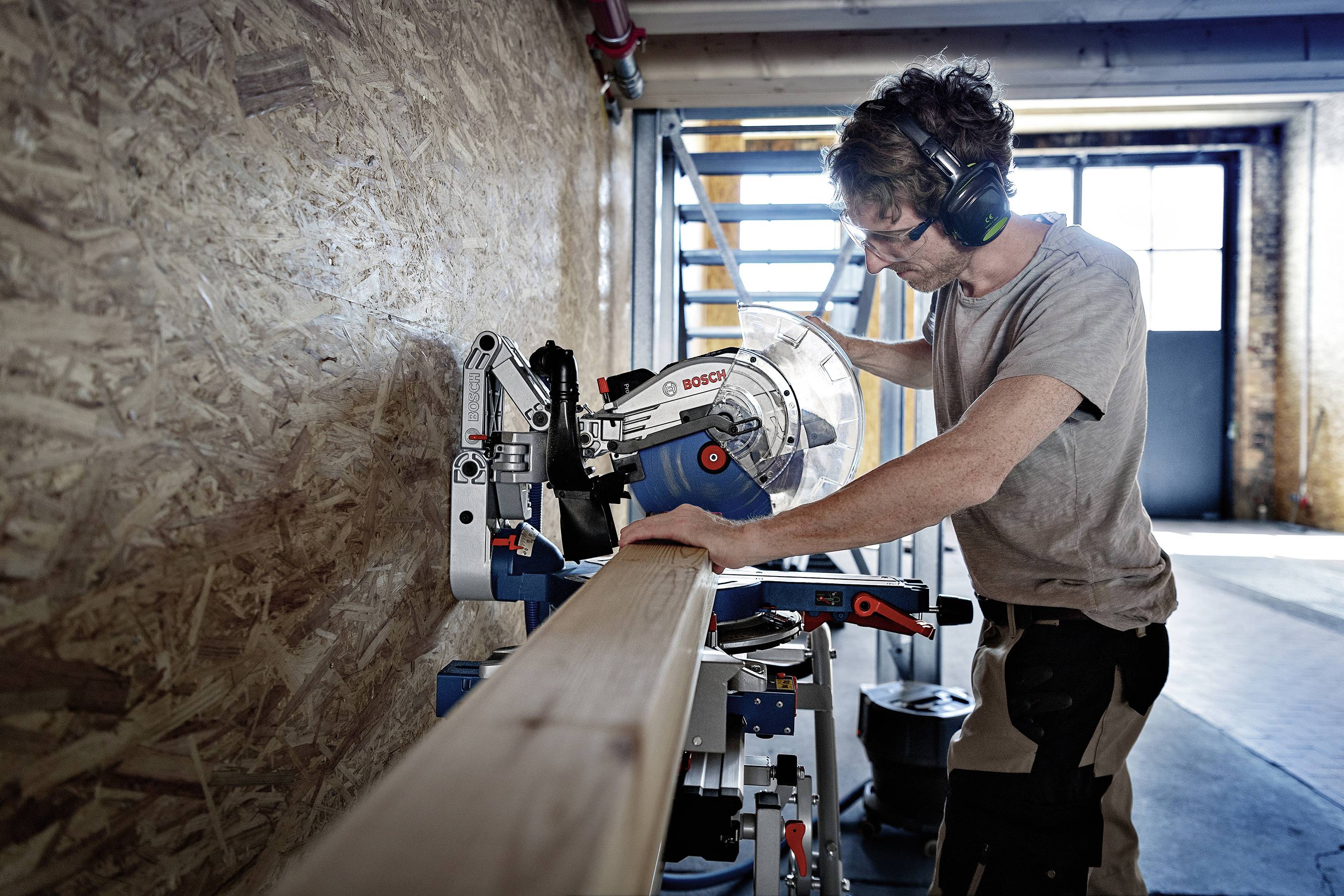 Eine Person in einem Werkraum schneidet mit einer elektrischen Säge ein Holzbrett. Sie trägt Schutzbrille und Gehörschutz.