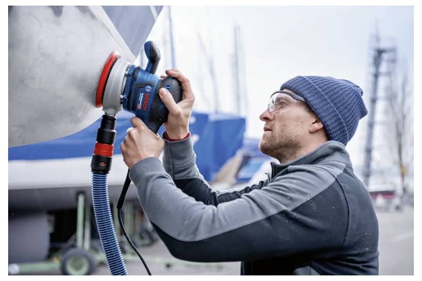 Eine Person mit Mütze benutzt ein Elektrowerkzeug, um den Rumpf eines Bootes in einem Yachthafen zu polieren. Im unscharfen Hintergrund sind Boote und Masten zu sehen.