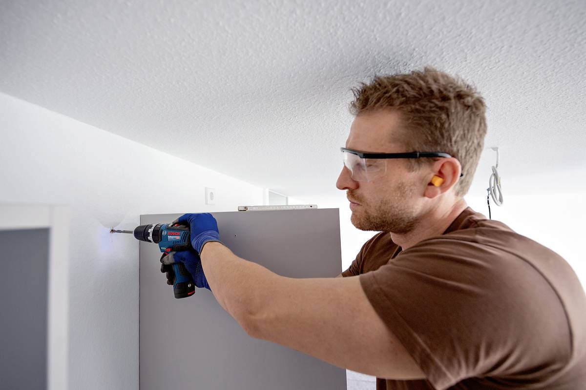 Ein Mann mit Schutzbrille und Gehörschutz bohrt ein Loch in die Wand. Er trägt blaue Handschuhe und verwendet eine elektrische Bohrmaschine.