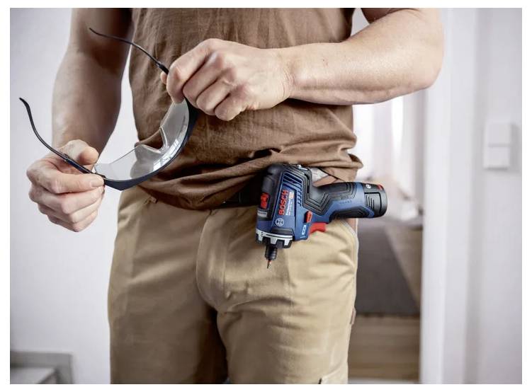 Eine Person in Arbeitskleidung hält eine Schutzbrille in der Hand, während ein Akkubohrer in einer vorderen Tasche steckt.