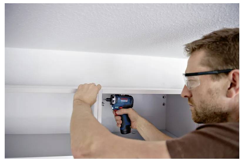 Ein Mann benutzt einen kabellosen Bohrer, um einen Schrank unter einem Regal zu installieren oder zu reparieren, Profilansicht in Innenräumen.