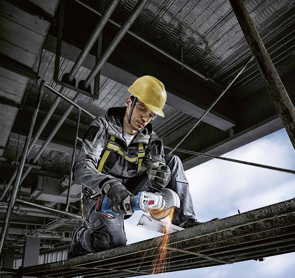 Ein Bauarbeiter mit Schutzhelm schneidet ein Metallrohr mit einem Winkelschleifer auf einer Baustelle. Funken fliegen vom Schneidpunkt.