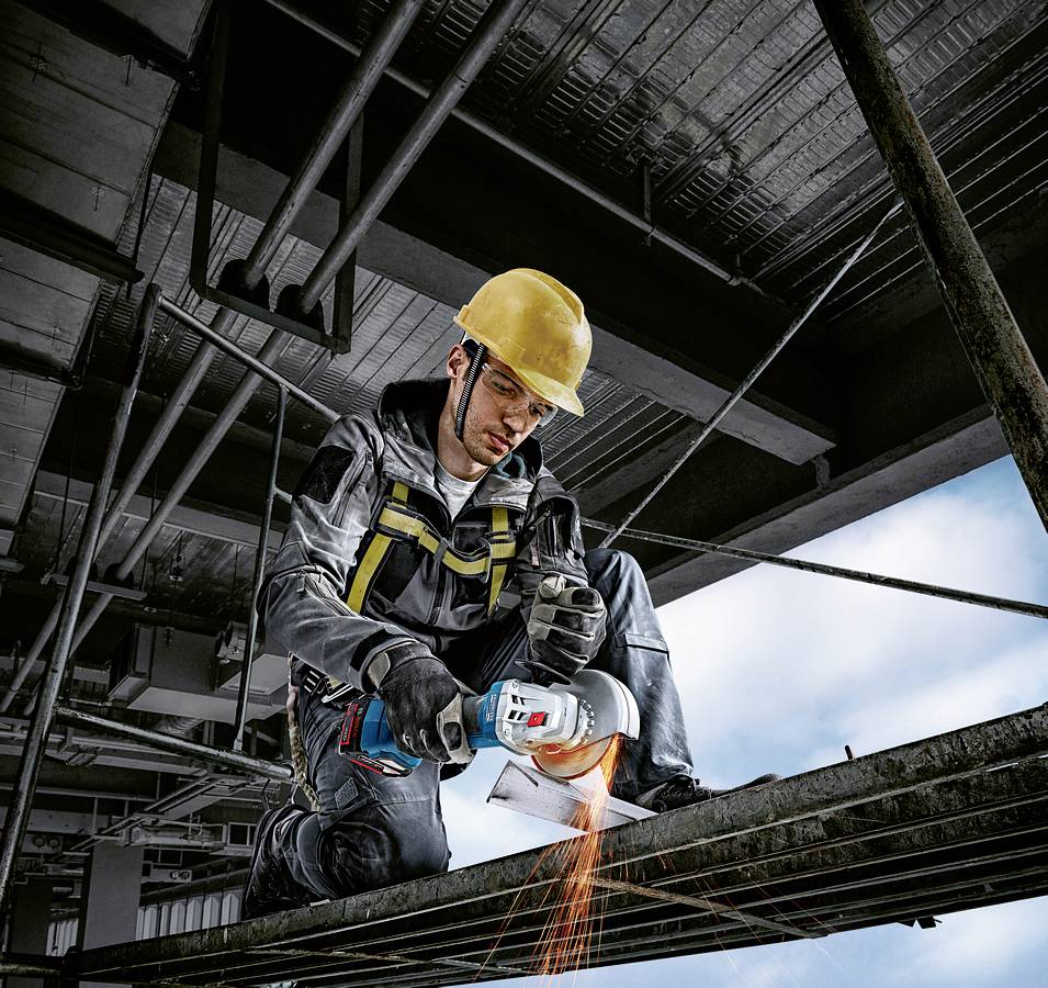 Ein Bauarbeiter mit Schutzhelm verwendet ein Elektrowerkzeug, um Metall zu schneiden. Funken fliegen, während er auf einem Stahlträger kniet.