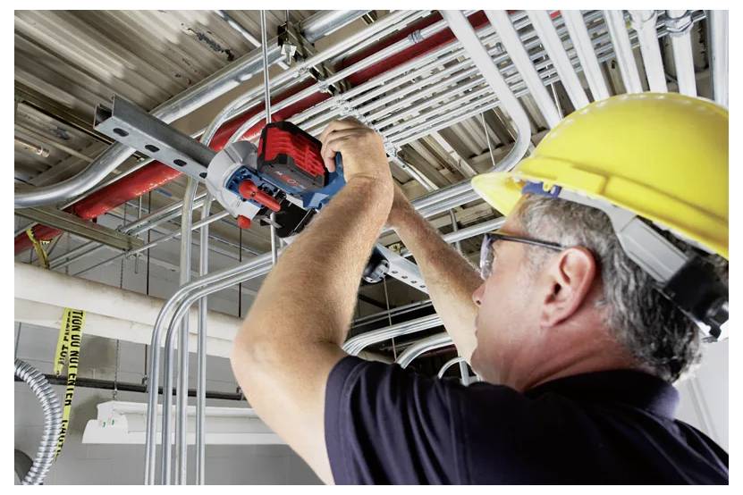 Arbeiter auf dem Bau mit gelbem Schutzhelm benutzt ein Elektrowerkzeug, um Stromleitung über Kopf in einem Deckengerüst zu installieren.