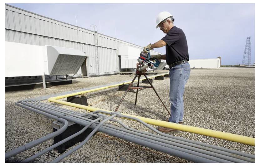 Bauarbeiter bohrt eine Leitung durch ein Rohrbündel auf einem Dach neben einem Industriegebäude.