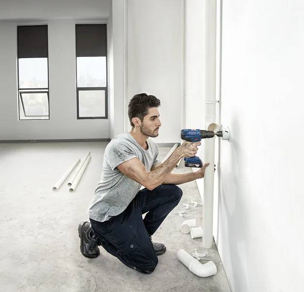 Eine Person kniet in einem unfertigen Raum und verwendet eine Bohrmaschine, um Rohre an einer weißen Wand zu installieren. Rohre und Fitting liegen auf dem Boden verstreut.