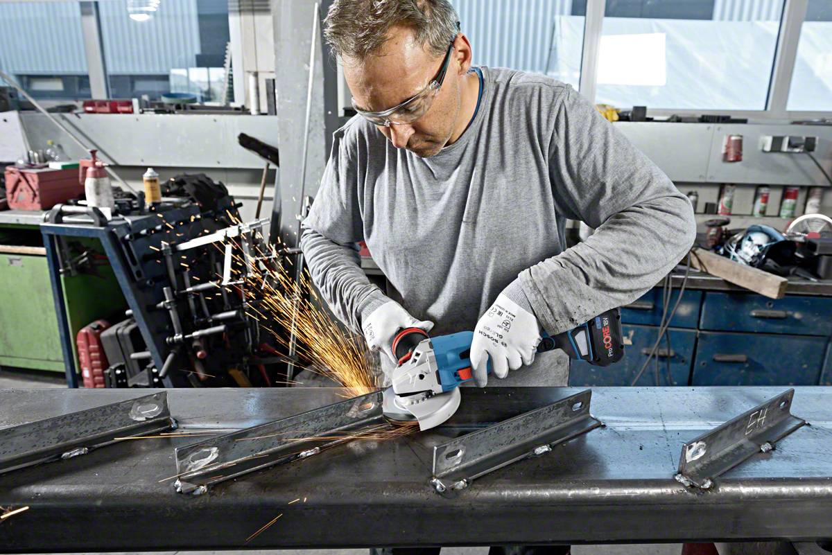 Ein Mann schleift Metall mit einem Winkelschleifer in einer Werkstatt. Funken fliegen, Schutzbrille und Handschuhe werden getragen.