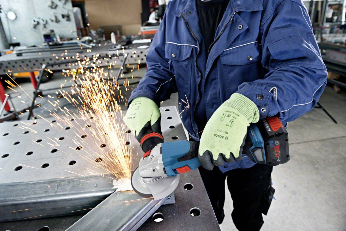 Eine Person schleift Metall mit einem Winkelschleifer, Funken sprühen. Die Person trägt Handschuhe und eine blaue Arbeitsjacke.