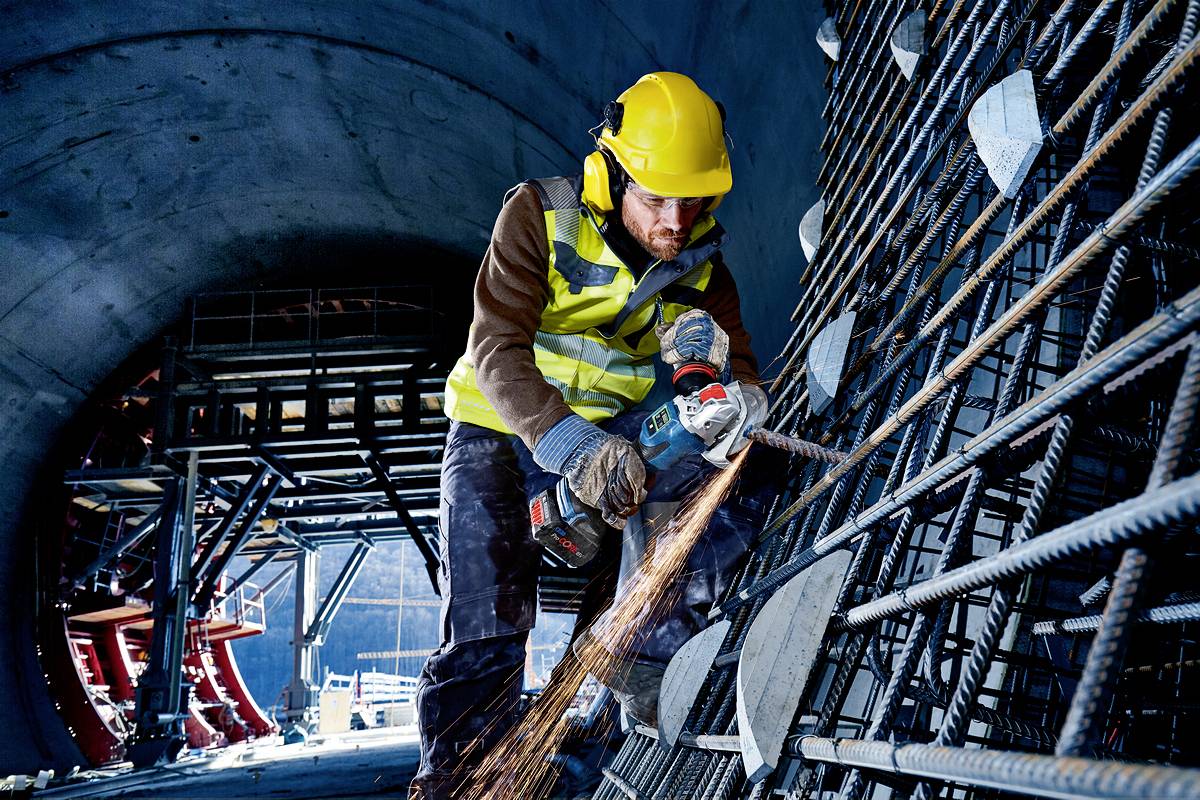 Ein Bauarbeiter schneidet mit einem Winkelschleifer Metallstäbe in einem Tunnel. Funken fliegen, er trägt Schutzkleidung und Helm.