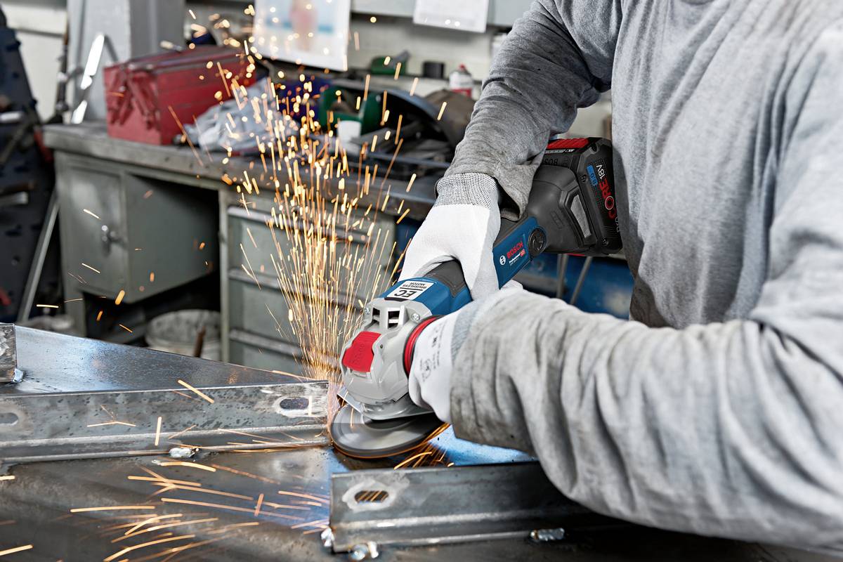 Eine Person schleift Metall mit einem Winkelschleifer in einer Werkstatt. Funken fliegen vom Metall weg, Arbeitsschutzkleidung wird getragen.
