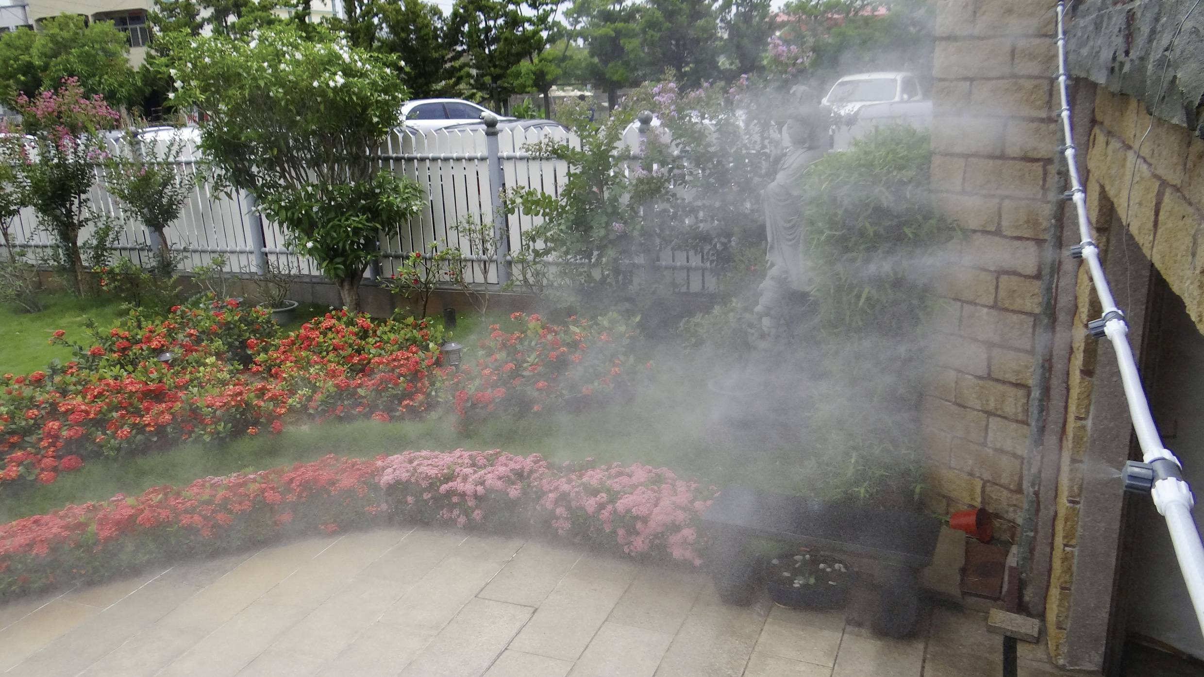 Ein Garten mit blühenden roten Blumen und grünen Büschen, bewässert durch einen feinen Wassernebel, der vor einer Steinmauer schwebt.