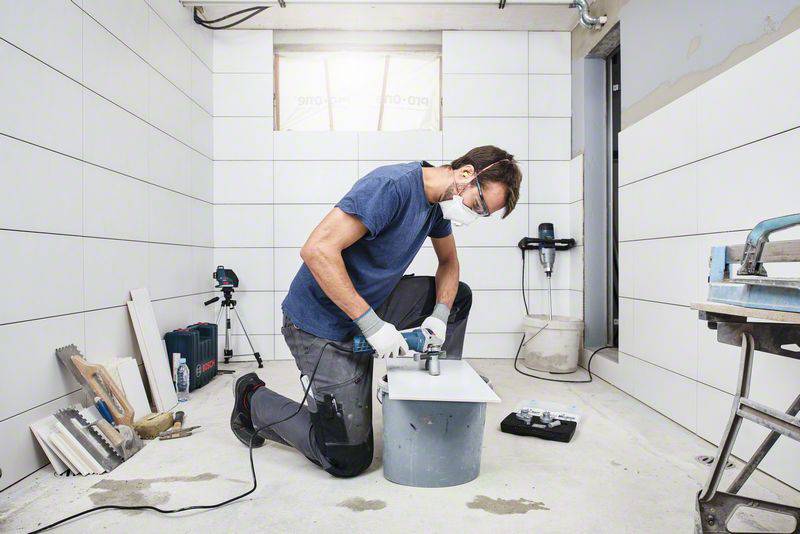 Eine Person mit Schutzmaske arbeitet in einem gefliesten Raum an einem Eimer. Auf dem Boden liegen Werkzeuge und Fliesen verteilt.