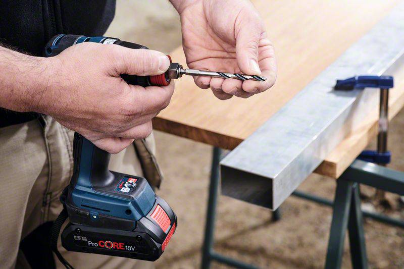 Eine Person hält eine Bohrmaschine und setzt einen Bohrer ein. Auf einem Tisch liegt ein Metallstück, das mit einer Klemme befestigt ist.