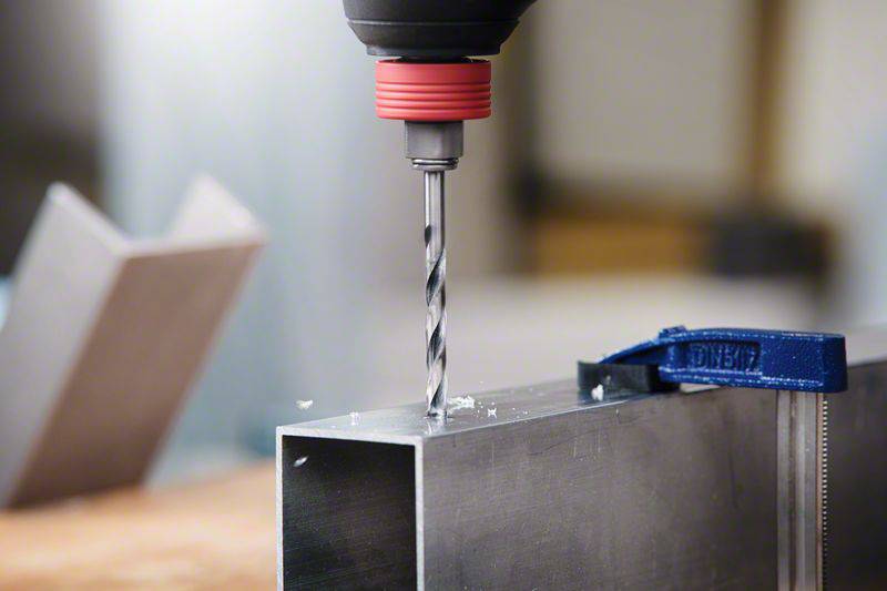 Eine Bohrmaschine bohrt ein Loch in ein rechteckiges Metallrohr, das auf einem Tisch befestigt ist; Späne fliegen weg.
